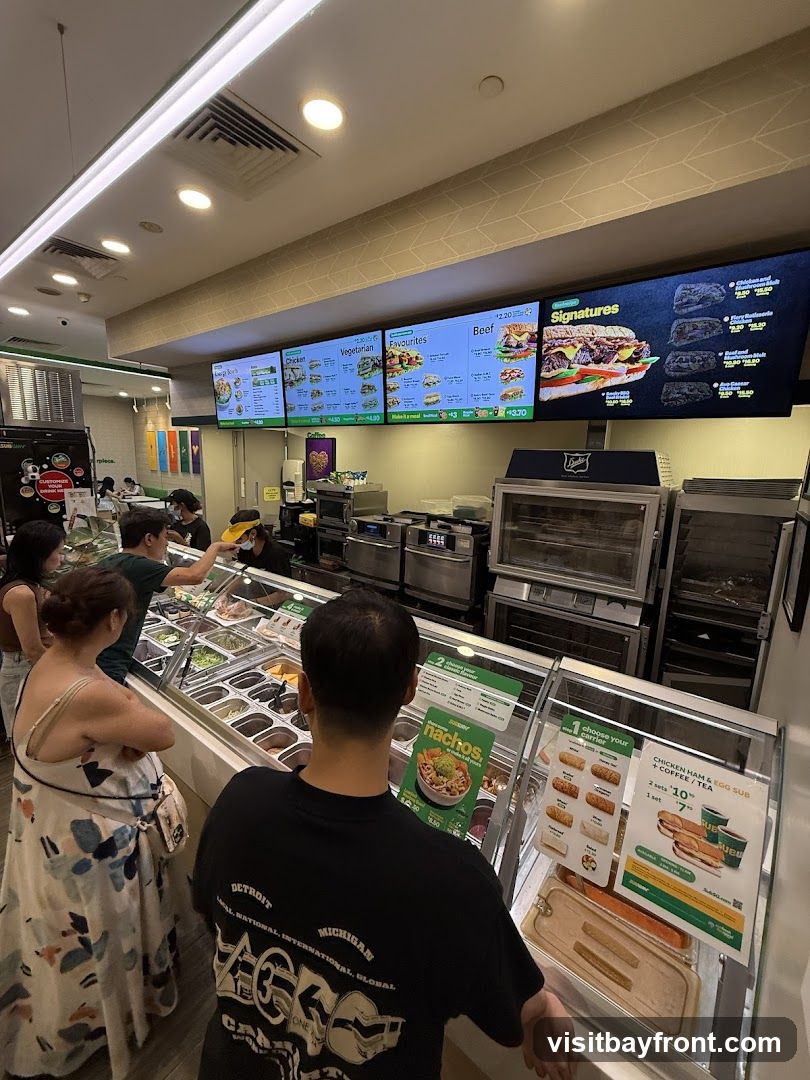 Customers ordering at a Subway restaurant. Digital menu boards display various sandwich options, including Chicken, Vegetarian, Beef, and Signatures. A display case shows fresh ingredients, and promotional materials for nachos and breakfast subs are visible.