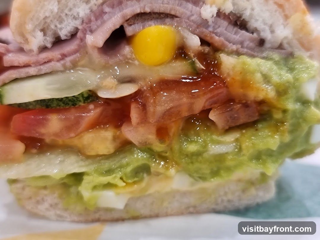 A close-up, slightly blurry image of a sandwich cut in half, revealing its layers. The sandwich appears to be made with a white roll and filled with sliced ham, corn, cucumber, tomato, lettuce, guacamole, and possibly chopped egg. A glossy sauce or dressing is also visible. The sandwich is presented on a surface with a green and white striped pattern.