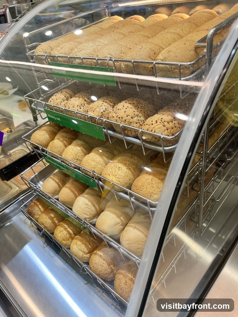 A refrigerated display case filled with various types of bread loaves and rolls. The breads are arranged on metal racks, with some loaves on the top shelf and smaller rolls on the lower shelves. Green labels indicate different bread varieties such as "Multigrain" and "Honey Oat." The display is clean and well-lit, showcasing the freshness of the baked goods.