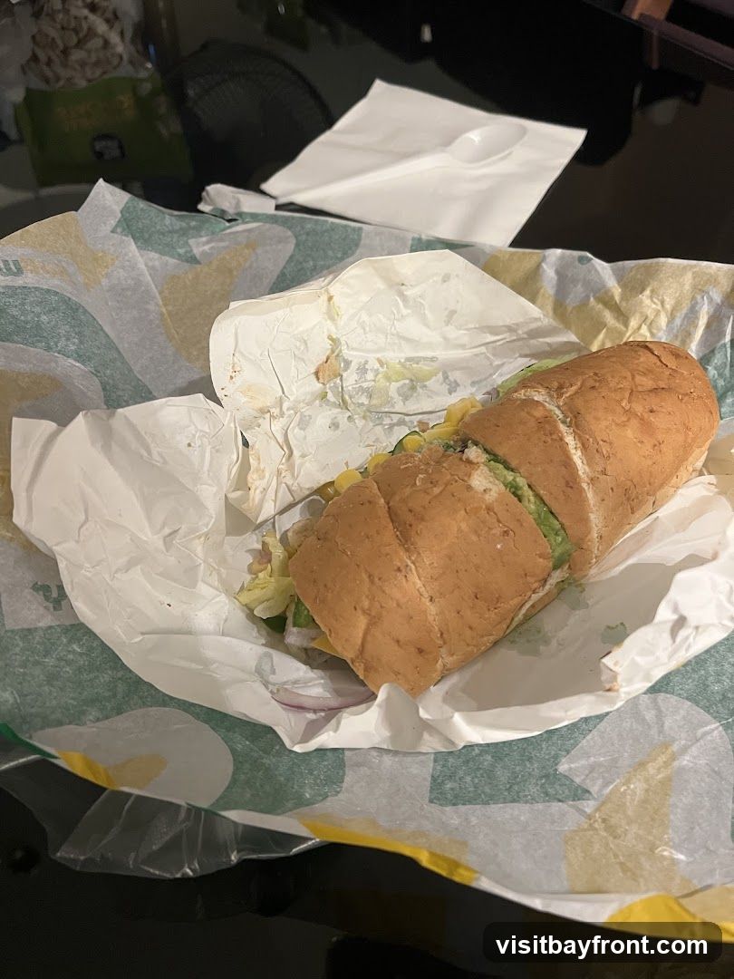 A half-eaten Subway sandwich is wrapped in white paper and placed on a green and white patterned paper liner. The sandwich appears to be on a whole wheat bun and contains lettuce, red onion, and avocado. A white plastic spoon and napkin are visible in the background.