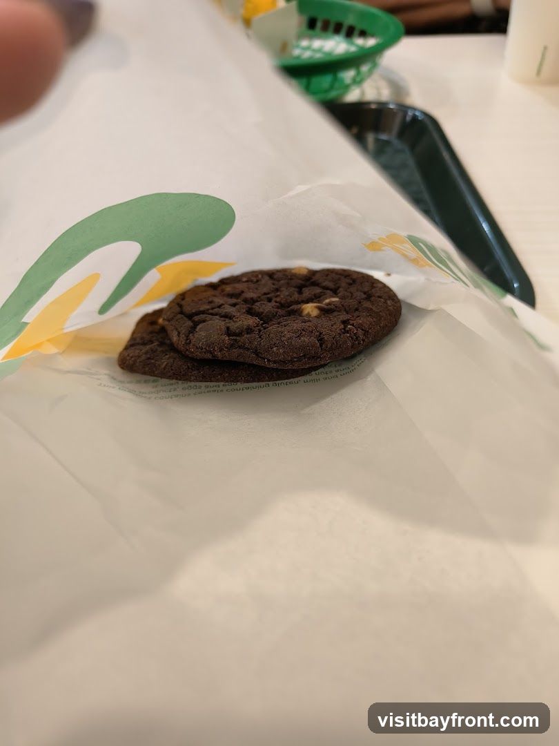 Two chocolate chip cookies are placed on white paper with a green and yellow logo. In the background, a green basket and a dark tray are visible.