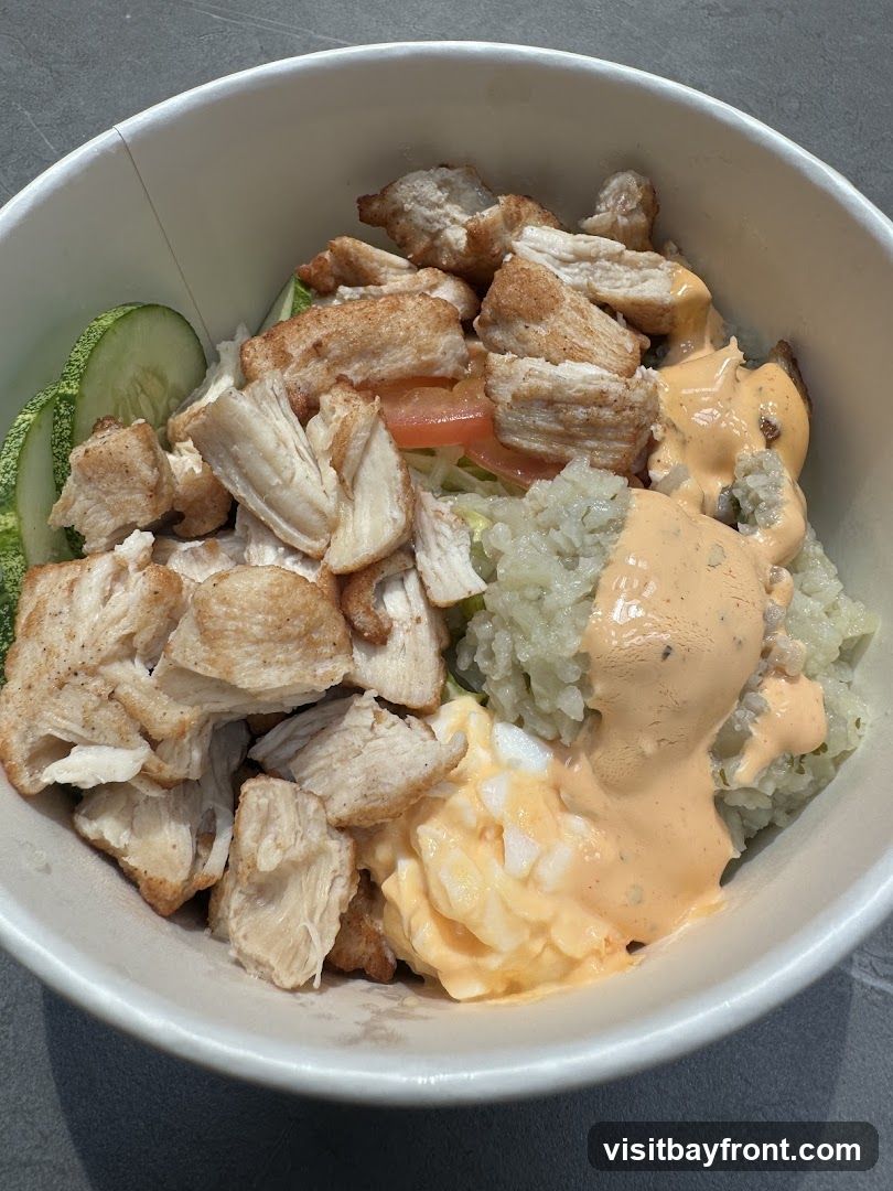 A close-up shot of a take-out bowl filled with food. The bowl contains chopped grilled chicken, rice, sliced cucumbers, chopped tomatoes, and a creamy orange sauce drizzled over the rice. There is also a portion of chopped hard-boiled eggs mixed with a similar sauce. The food is presented in a white paper bowl with a slightly textured exterior.