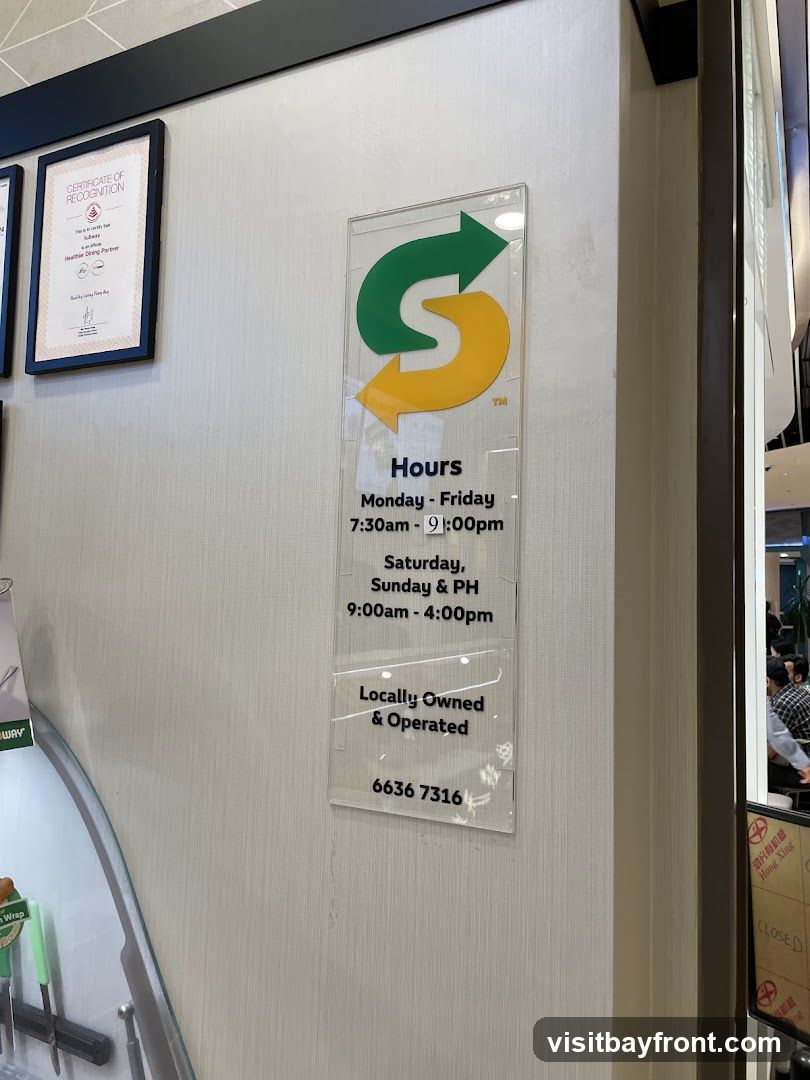 A clear acrylic sign displaying the Subway logo and operating hours. The sign indicates that the establishment is open Monday to Friday from 7:30 AM to 9:00 PM, and on Saturdays, Sundays, and public holidays from 9:00 AM to 4:00 PM. It also states that the business is locally owned and operated and provides a contact number. A certificate of recognition is also visible on the wall.