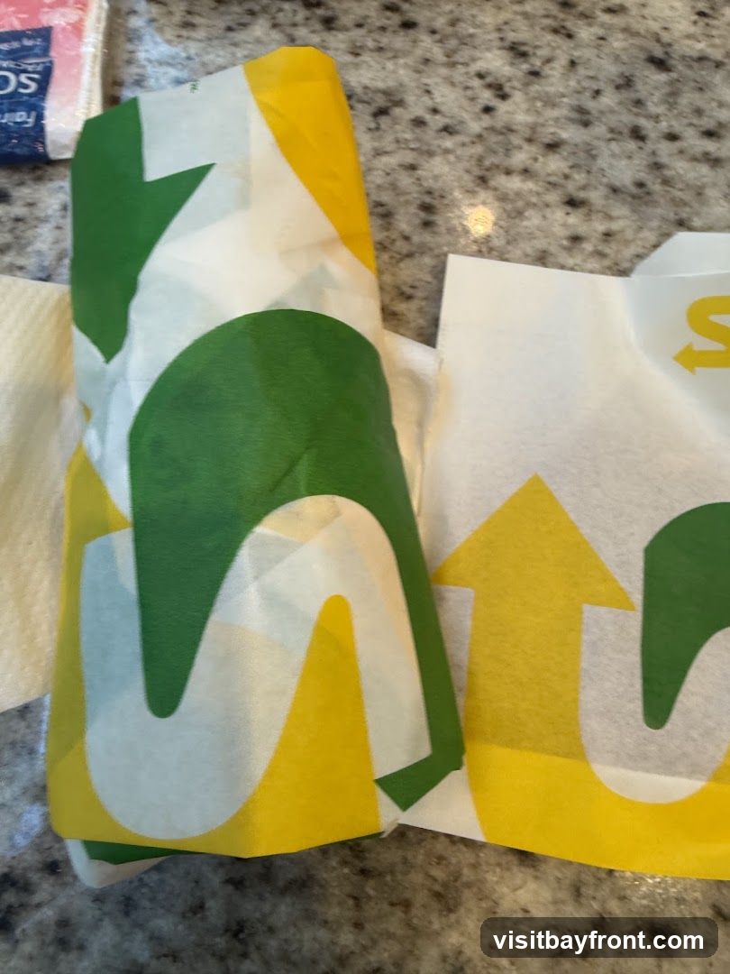 A sandwich wrapped in branded Subway paper. The paper is white with green and yellow arrows and the Subway logo.