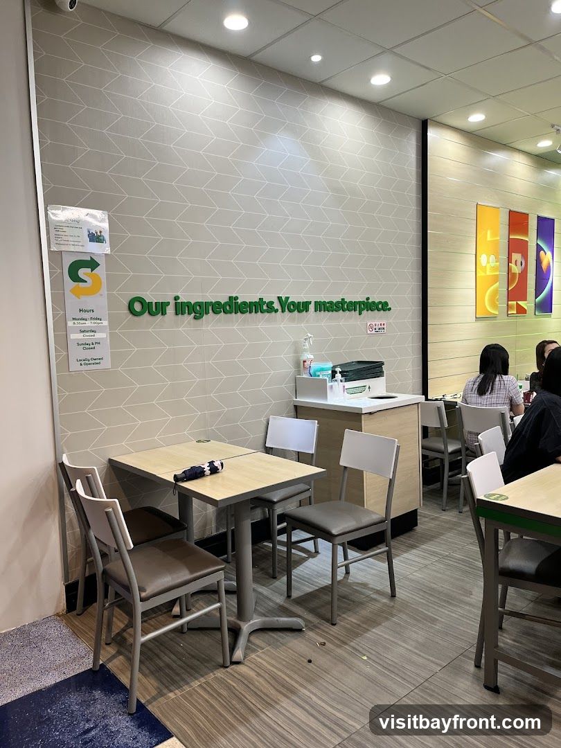 The interior of a Subway restaurant with tables and chairs. A sign on the wall reads "Our ingredients. Your masterpiece." and displays the Subway logo and hours of operation. Several people are seated at tables in the background.