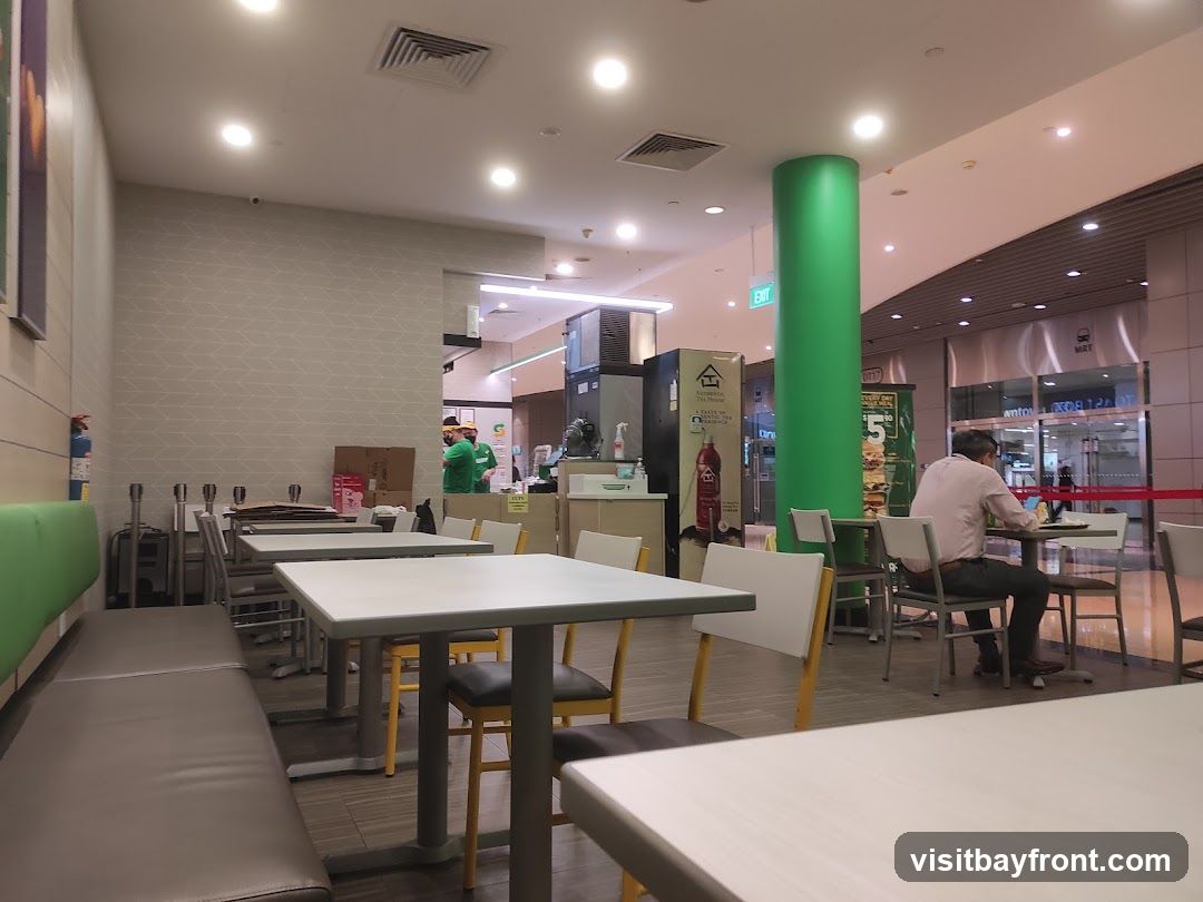 The image shows the interior of a fast-food restaurant with tables and chairs. A man is seated at a table in the background, and two staff members in green uniforms are visible behind the counter. A large green pillar is prominent in the center of the image. There are also promotional posters and signage visible.
