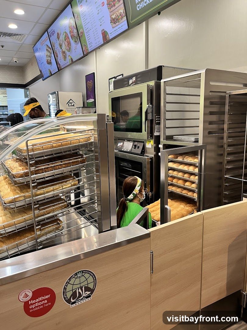 A view inside a bakery with trays of donuts and bread. A person in a green shirt is seen placing a tray of bread into an oven. Digital menus are displayed on the wall above. A Halal certification sticker is visible on the counter.