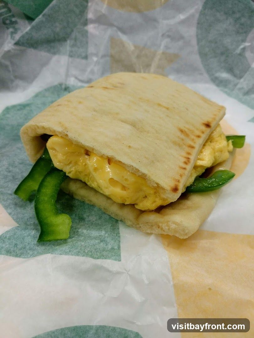 A breakfast sandwich made with flatbread, scrambled eggs, and green bell peppers, wrapped in branded paper.