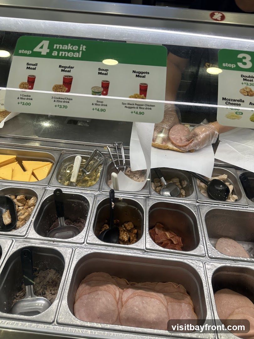 A display of ingredients and meal deal options at a Subway restaurant. The image shows various meats, cheeses, and toppings in metal containers. A sign above details "step 4 make it a meal" with options like Small Meal, Regular Meal, Soup Meal, and Nuggets Meal, along with their prices and included items. Another sign indicates "step 3" with options like Double Meat and Mozzarella Cheese. A hand in a glove is seen preparing a sandwich.