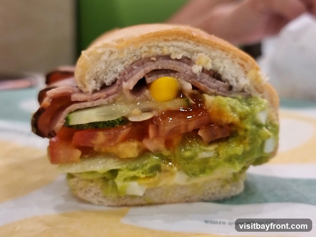 A close-up, slightly angled shot of a sandwich cut in half, revealing layers of ham, cucumber, tomatoes, lettuce, corn, and guacamole. The sandwich is on a bun and appears to be served on a patterned paper liner.