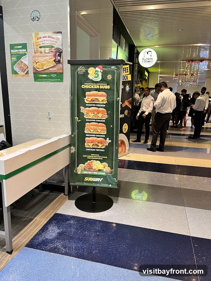 A promotional banner for Subway's chicken subs is displayed in front of a counter. The banner highlights various chicken subs including Roasted Chicken, Bulgogi Chicken, Breaded Chicken, and Chicken Teriyaki. It also mentions a limited-time offer with a mint chocolate chip cookie and chicken bowl. In the background, other people are visible, and a sign for Guzman Y Gomez can be seen.