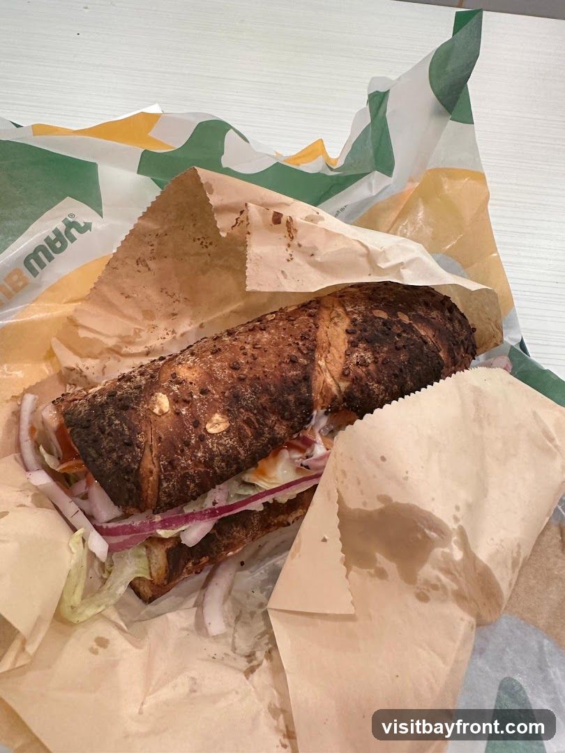 A close-up, overhead view of a Subway sandwich wrapped in branded paper. The sandwich is on a dark, seeded bread and filled with lettuce, red onion, and sauce. The paper wrapping features the Subway logo and colors.