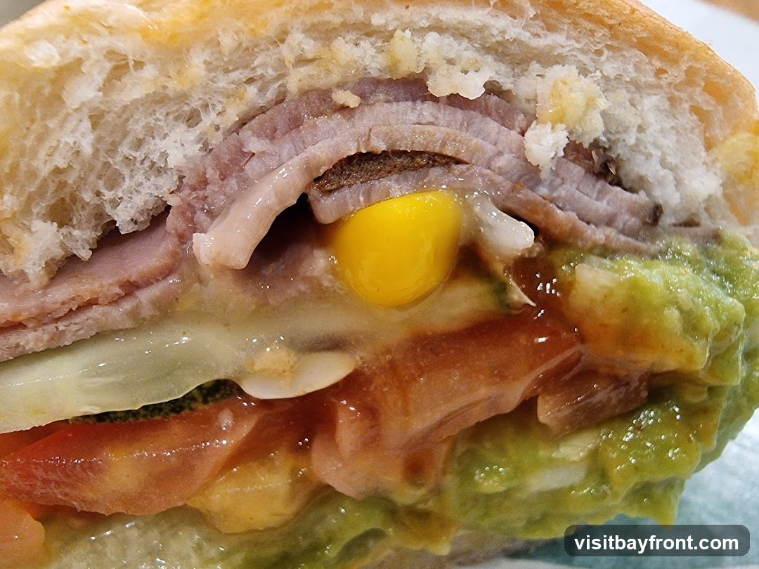 A close-up shot of a sandwich reveals layers of thinly sliced meat, possibly ham, stacked on a soft white bun. Visible ingredients include a bright yellow corn kernel, a dollop of green guacamole, sliced tomatoes, and what appears to be melted cheese. The texture of the bun is airy and slightly toasted, while the fillings are moist and colorful.