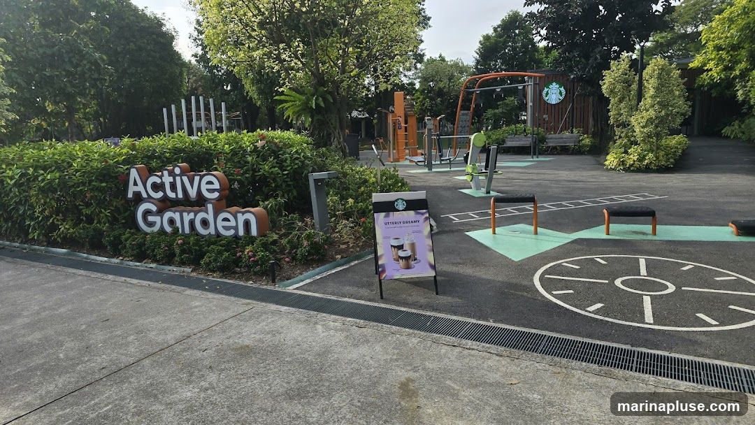 An outdoor fitness area with exercise equipment and a Starbucks advertisement. The advertisement is an A-frame sign displaying a promotional image of two iced coffee drinks with the text "UTTERLY DREAMY" and "Escape to your happy place". The Starbucks logo is visible on the sign and on a wooden fence in the background. The area is surrounded by lush greenery and trees.