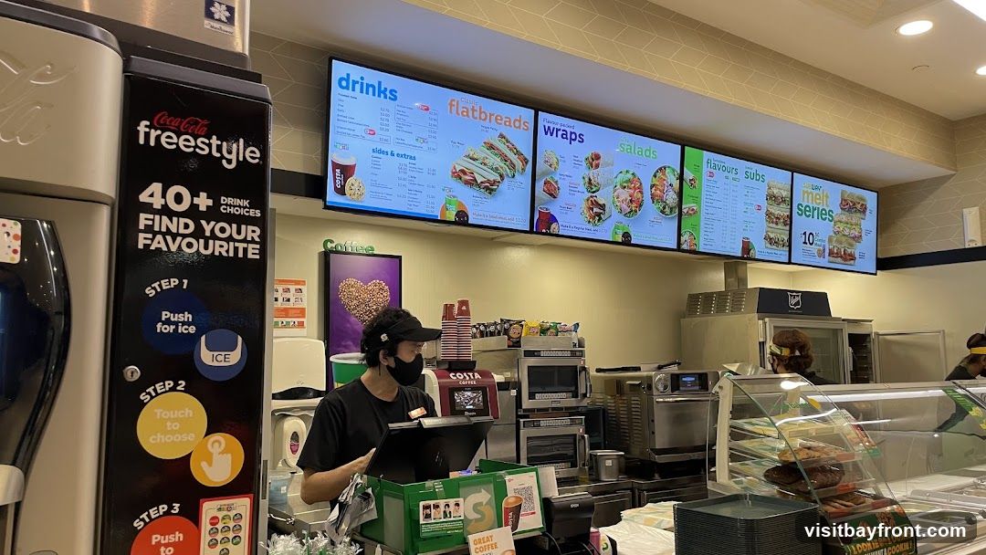 A view of a fast food restaurant counter with digital menus displaying drinks, flatbreads, wraps, salads, and subs. A Coca-Cola Freestyle machine is visible on the left, and a staff member wearing a mask and visor is serving customers. The interior features a clean, modern design with bright lighting.