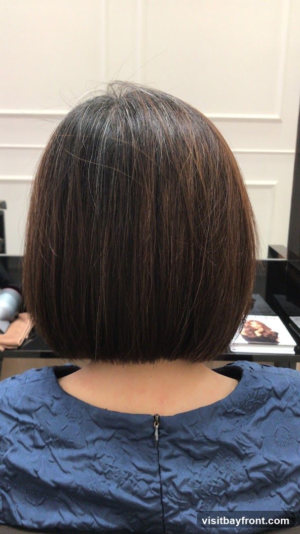 A person's back is shown, featuring a neatly cut bob haircut. The hair is dark brown with visible streaks of gray, particularly noticeable at the crown. The person is wearing a textured blue dress with a zipper closure at the back. The background appears to be an indoor setting, possibly a salon, with a black countertop and some items on it, including what looks like a magazine or book with an image of hair on the cover.