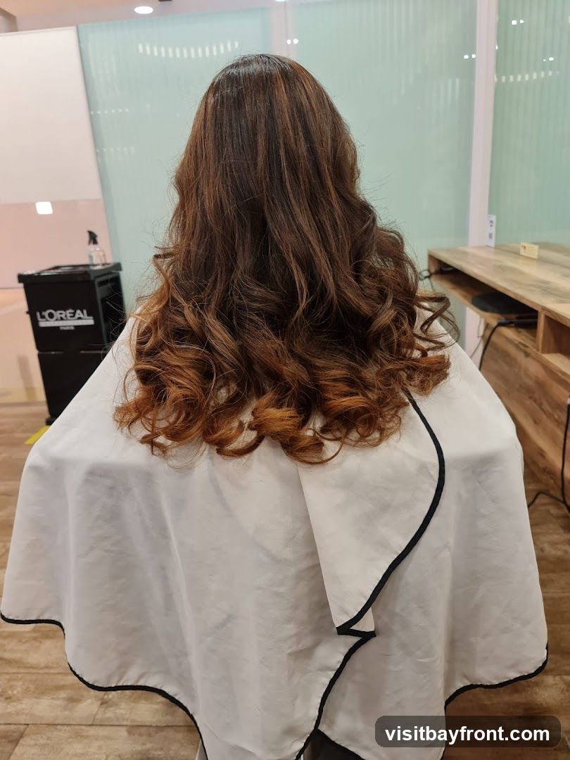 A woman's back is shown, with her long, wavy, brown hair styled with curls. She is wearing a white cape with black trim, commonly used in hair salons. In the background, a black trolley with the L'Oréal logo is visible, along with a wooden counter and a blurred interior.