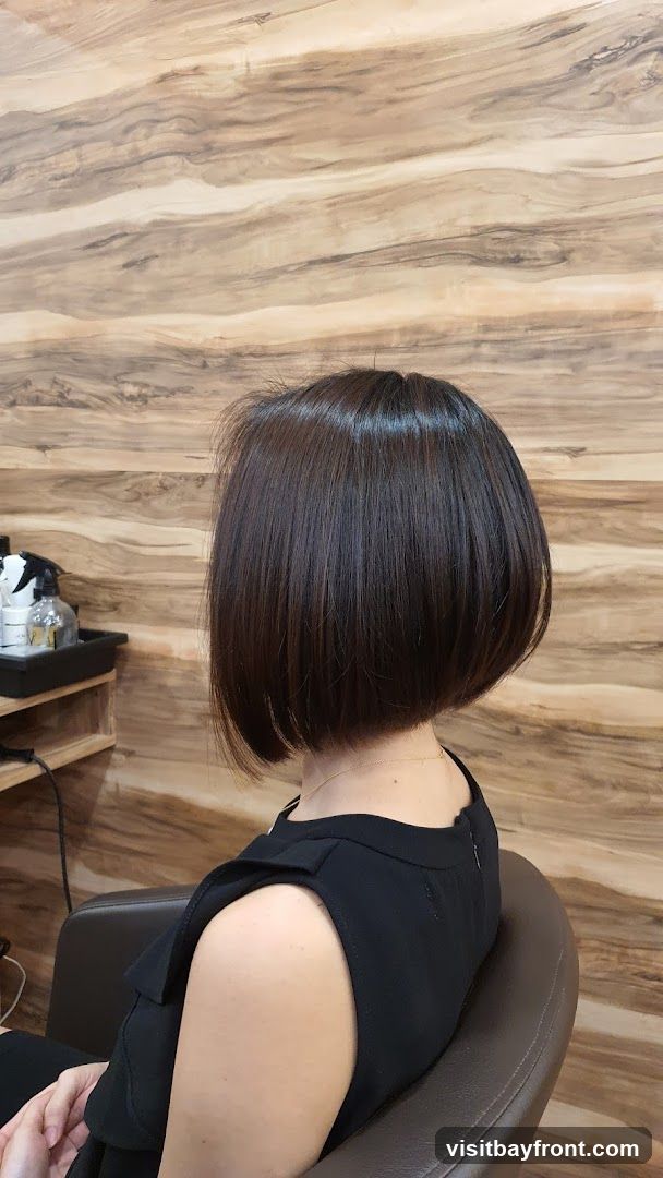A woman with a dark brown bob haircut is sitting in a salon chair. The background features a wood-paneled wall. To the left, a shelf holds bottles and a spray bottle, suggesting a hair salon environment.