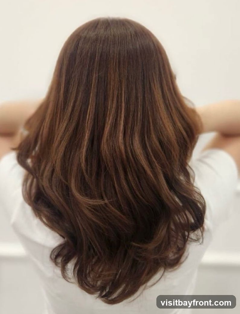A close-up shot from behind of a woman with long, wavy, dark brown hair with subtle highlights. She is wearing a white shirt and her hands are resting on the back of her head. The background is a plain, light color.