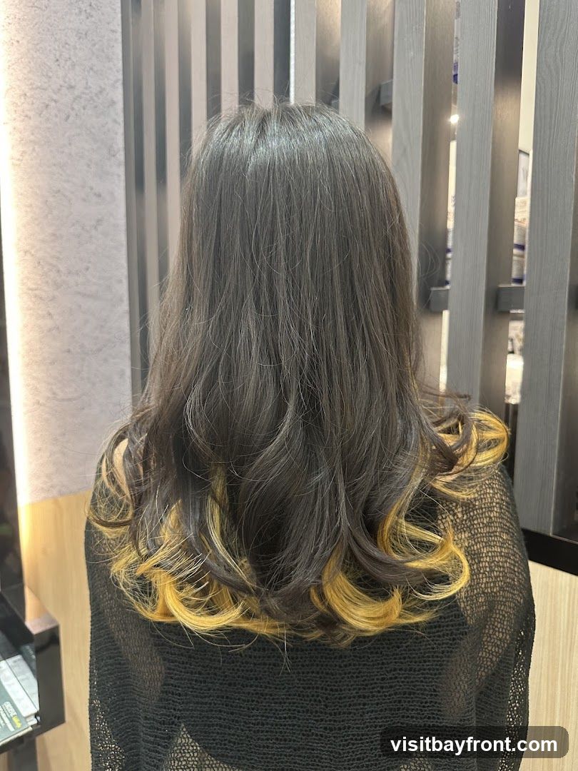 A woman's back is shown, displaying her dark, wavy hair with blonde highlights at the ends. She is wearing a black, sheer top. The background features vertical wooden panels and a blurred interior with shelves of products.