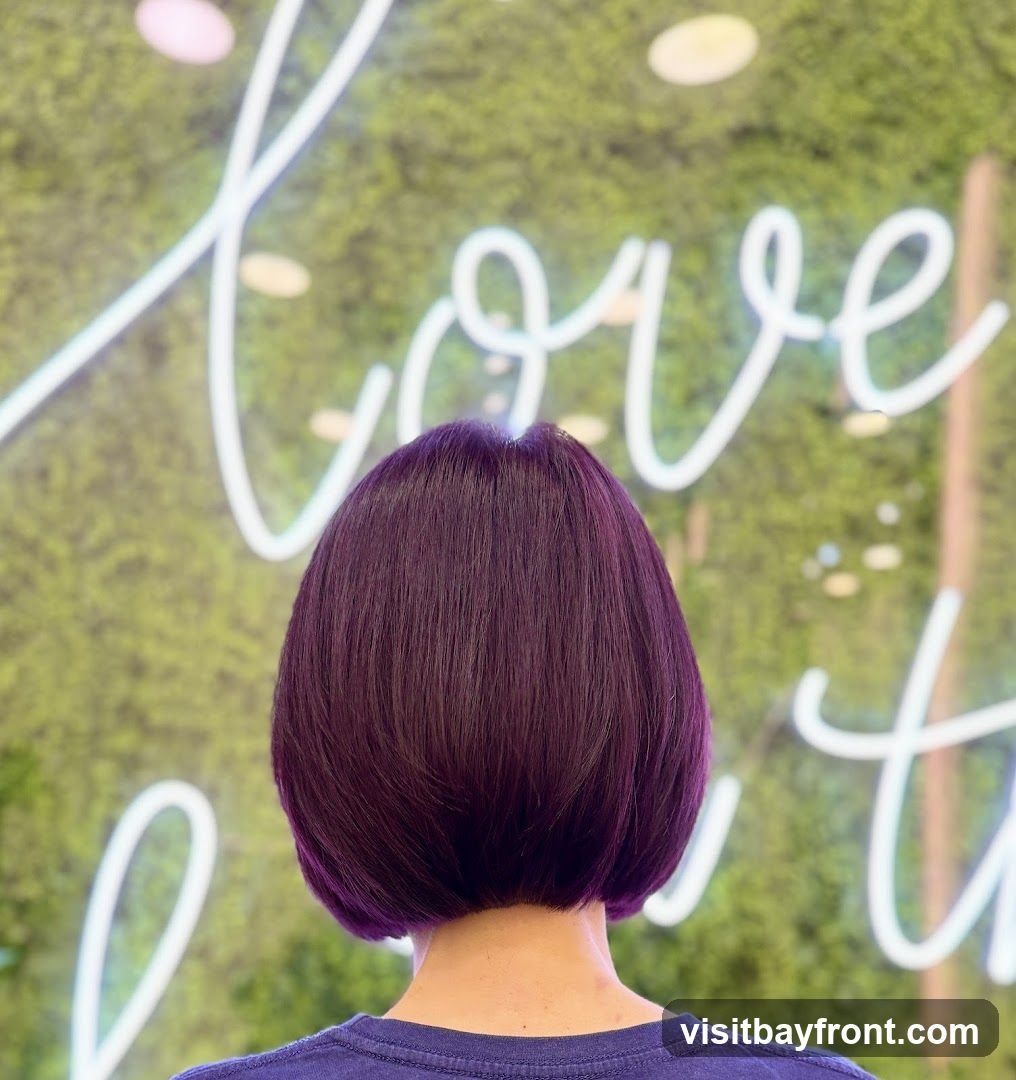 A person with a short, purple bob haircut is shown from behind, facing a wall with a green, moss-like texture. A white neon sign with cursive writing is visible on the wall. The writing appears to say "love" and possibly other words.
