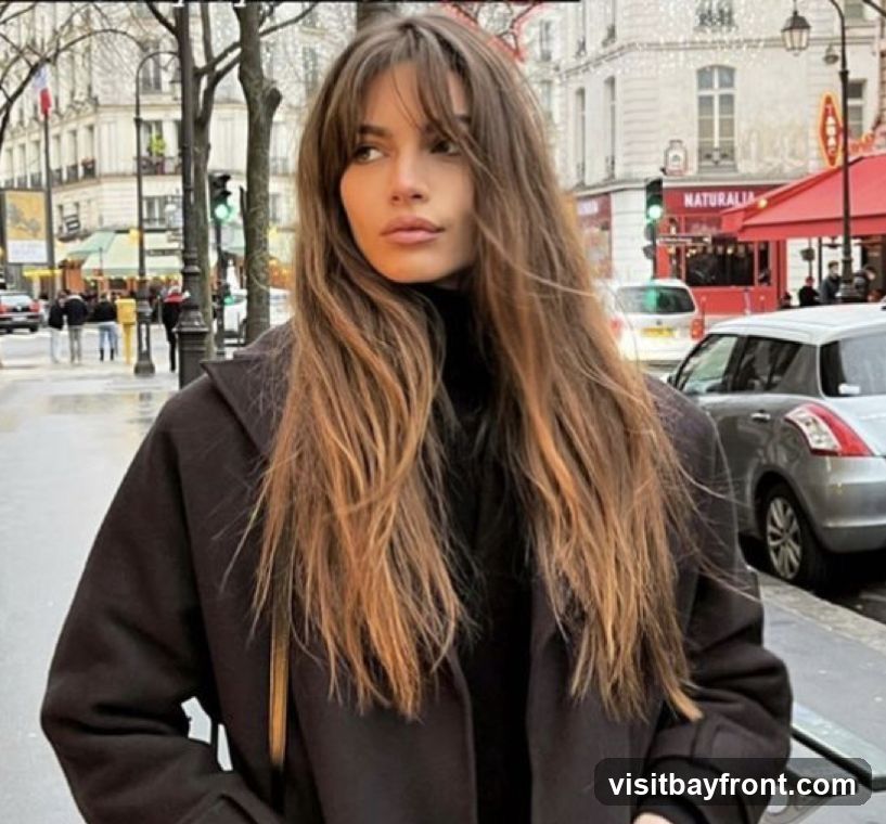A woman with long, wavy brown hair and bangs stands on a wet city street, looking to the side. She is wearing a dark turtleneck and a dark coat. In the background, there are buildings, cars, and streetlights. One building has a sign that reads "NATURALIA" and a red awning.