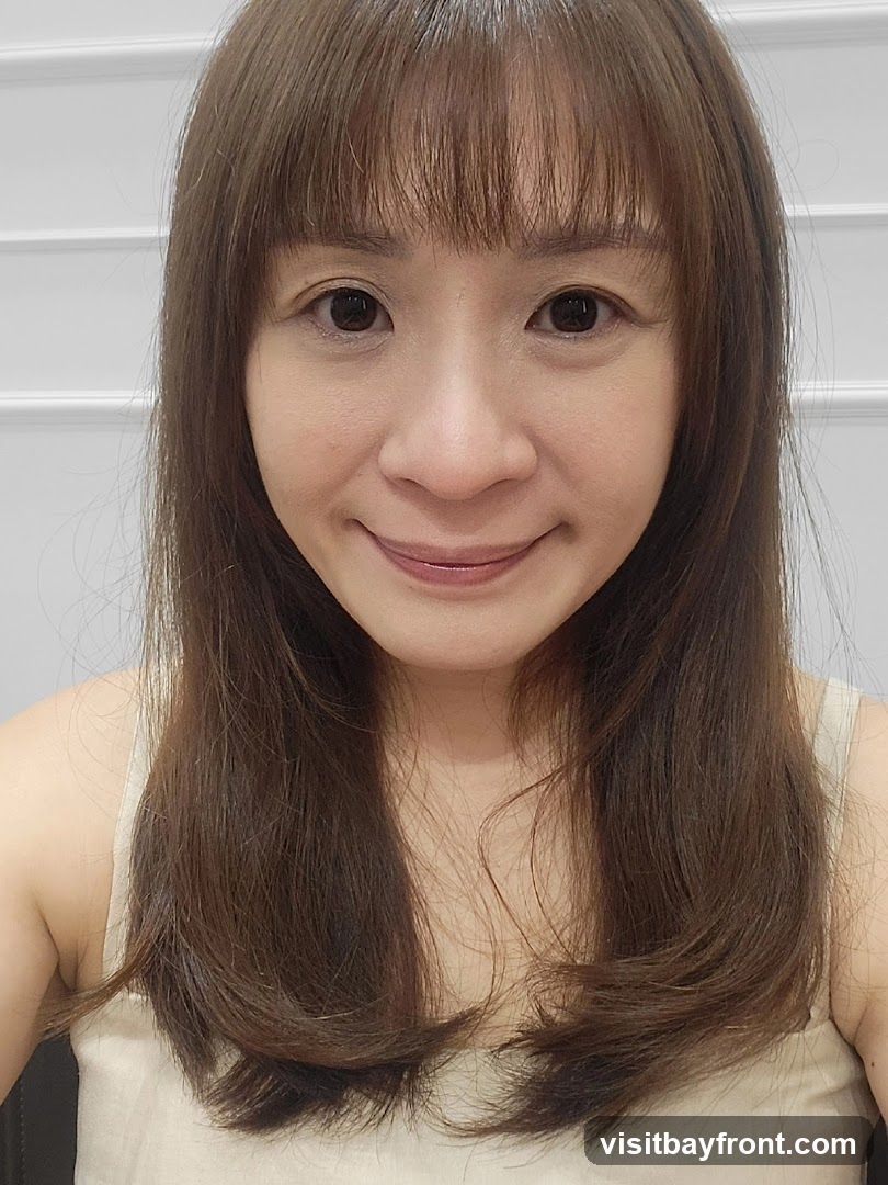 A close-up selfie of a woman with shoulder-length brown hair and bangs. She is wearing a light-colored tank top. The background is a white wall with horizontal paneling.