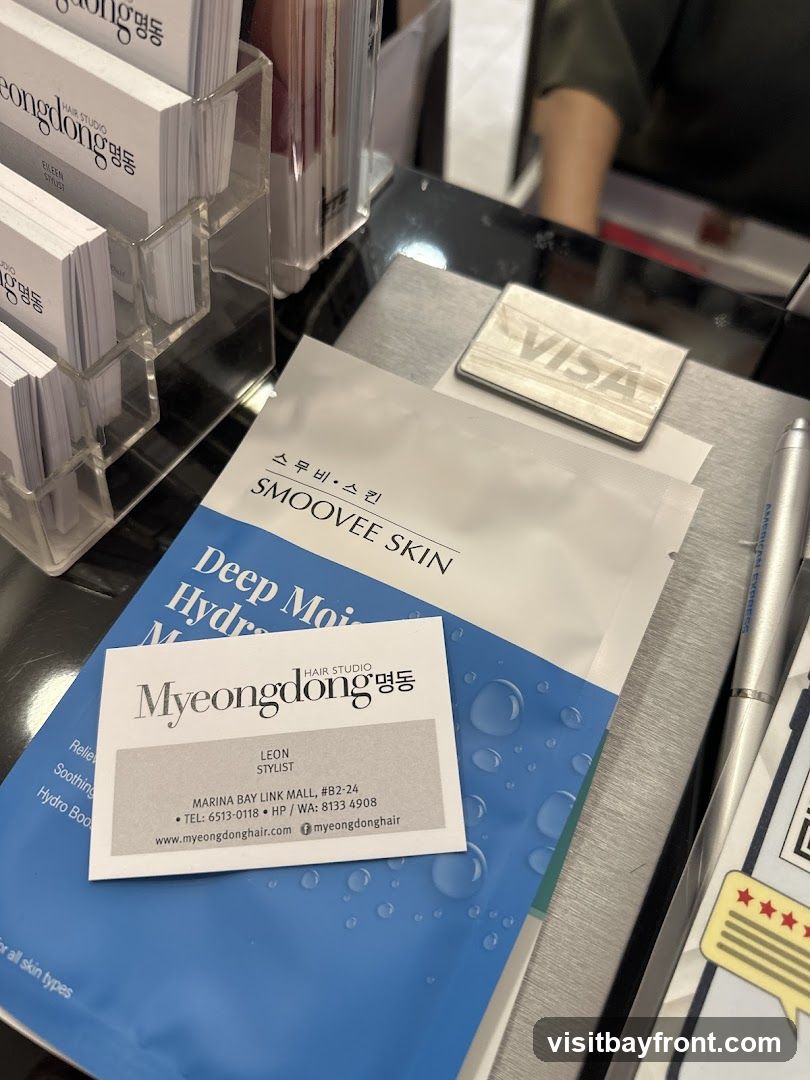 A close-up view of a counter at a hair studio, featuring business cards, a facial mask package, and a Visa card holder. The business cards are for Myeongdong Hair Studio and list contact information and website. The facial mask is labeled "SMOOVEE SKIN Deep Moisture Hydro Mask". A Visa card holder and a pen are also visible.