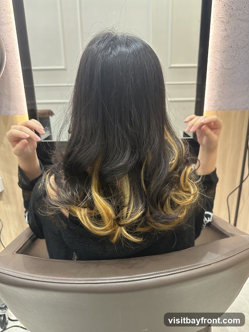 A woman is seated in a salon chair, viewed from behind. Her dark hair has been styled with loose curls and features prominent blonde highlights. She is wearing a black, sheer top. The background shows a mirror reflecting the interior of a salon, with clean, modern decor.