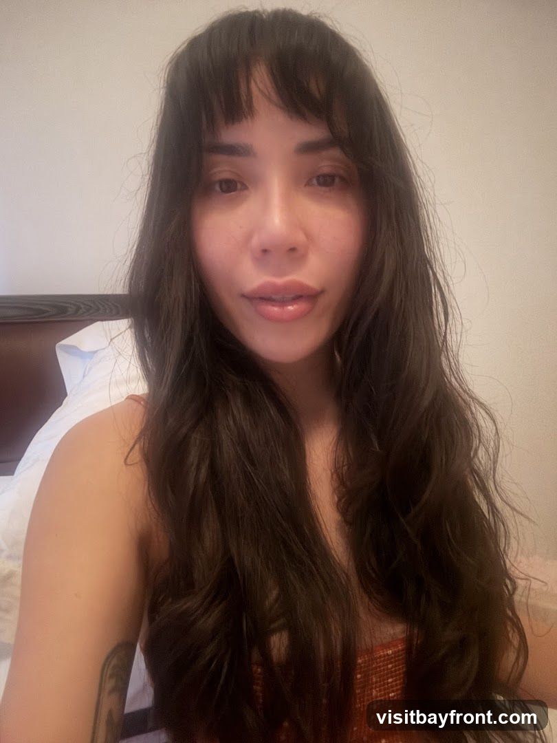 A close-up, head-and-shoulders shot of a woman with long, dark, wavy hair and bangs. She is looking directly at the camera. She is wearing a sparkly, orange top. The background is a plain wall and a pillow.
