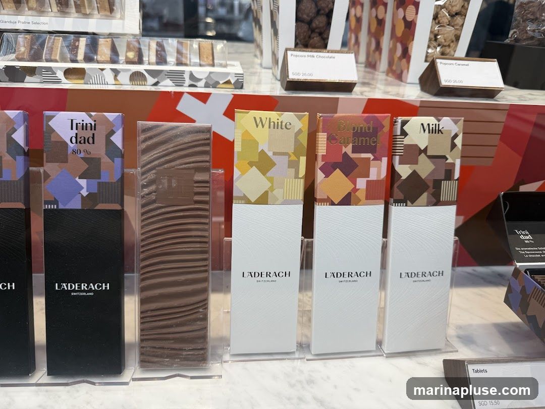 A display of various Laderach chocolates, including bars with different flavor labels like 'Trinidad 80%', 'White', 'Blond Caramel', and 'Milk'. There are also boxes of popcorn chocolates labeled 'Popcorn Milk Chocolate' and 'Popcorn Caramel', priced at SGD 26.00. A separate display shows 'Tablets' priced at SGD 15.50.