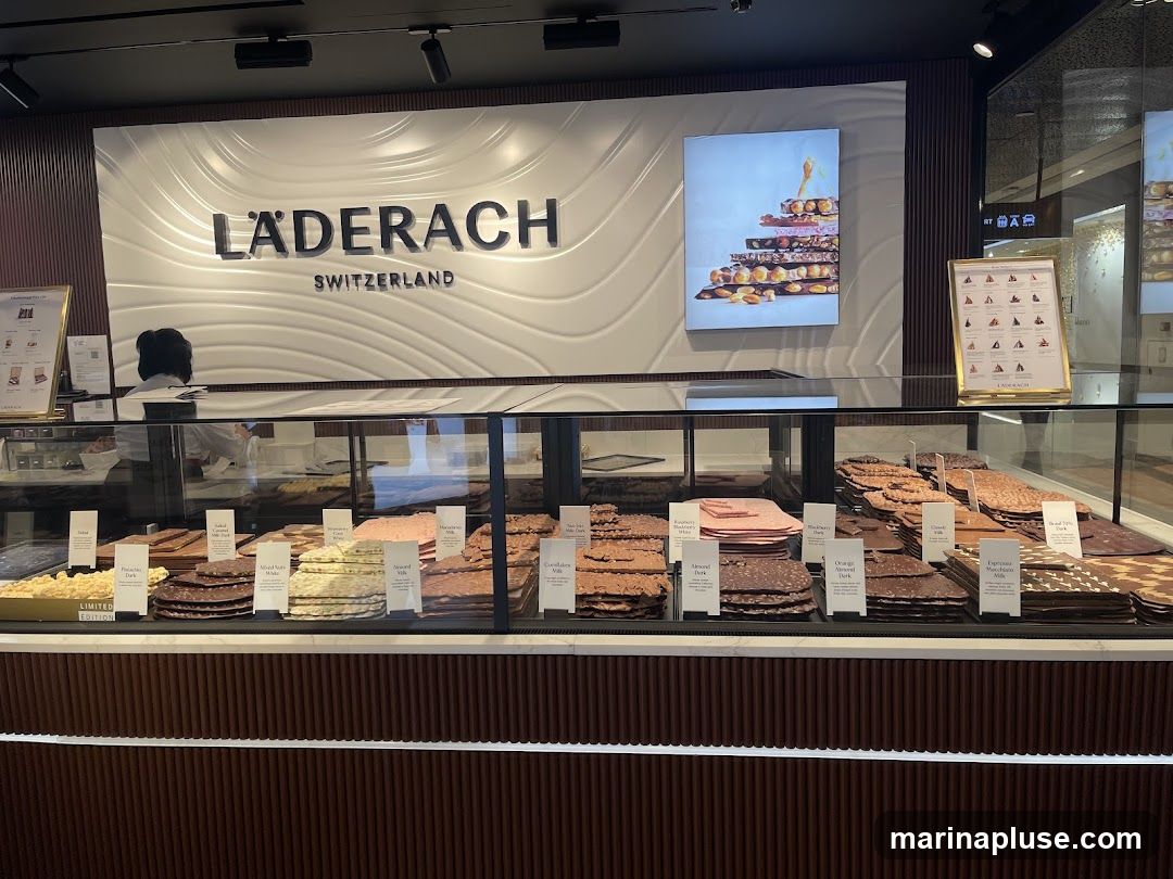 A display case filled with various types of chocolate bars and slabs, with the Läderach Switzerland logo prominently displayed on the wall above. There are also informational posters and a framed menu or product list.