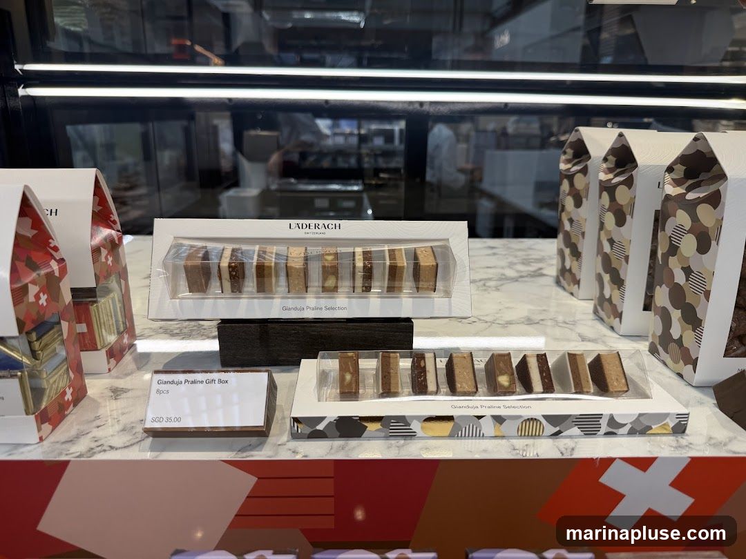 A display of Läderach chocolates, featuring several boxes of Gianduja Praline Selection and a Gianduja Praline Gift Box priced at SGD 35.00 for 8 pieces. The chocolates are arranged on a marble-patterned surface, with a Swiss flag motif visible at the bottom. In the background, a person in a white uniform is visible behind glass, possibly a staff member in the shop.