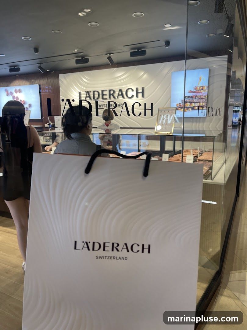 A shopping bag with the Laderach logo is held in the foreground, obscuring part of the shop entrance. Through the glass storefront, the Laderach logo is visible on the wall behind the counter. Display cases filled with chocolate products are visible, and staff members are attending to customers. A digital screen displays an image of a stacked chocolate creation.