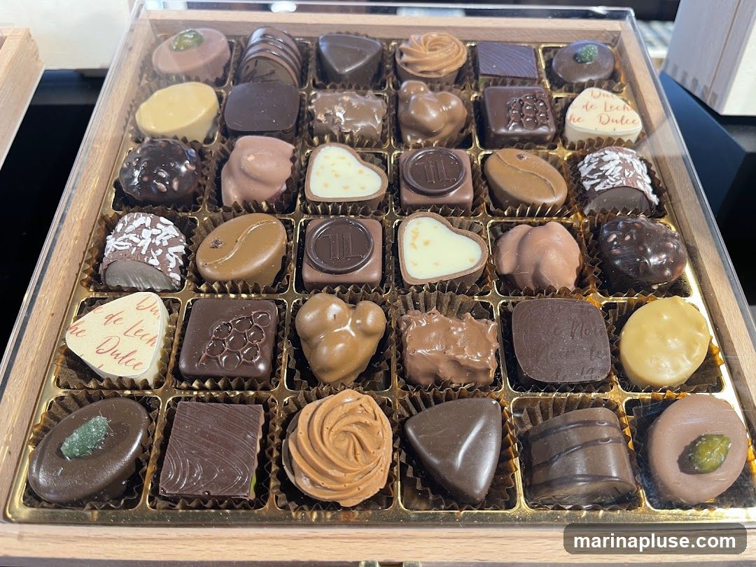 A close-up, overhead view of a wooden box filled with an assortment of chocolates. The chocolates vary in shape, size, and topping, with some appearing to be filled or coated. Several chocolates have decorative patterns or inscriptions. One chocolate has a green garnish on top. Another has a white heart shape with red text that reads "de Leche Dulce". The chocolates are arranged in a grid within the box.