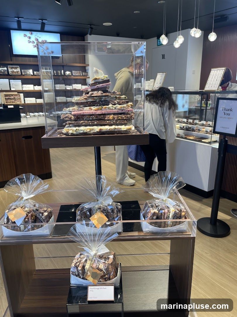 A view inside a modern chocolate shop. In the foreground, there are several individually wrapped chocolate gift packs displayed on shelves. A tall, tiered display of various chocolate bars is prominently featured in a glass case. In the background, customers are browsing at a counter with more chocolates on display, and shelves stocked with more chocolate boxes line the walls. A sign reads "Thank You, See you again!".