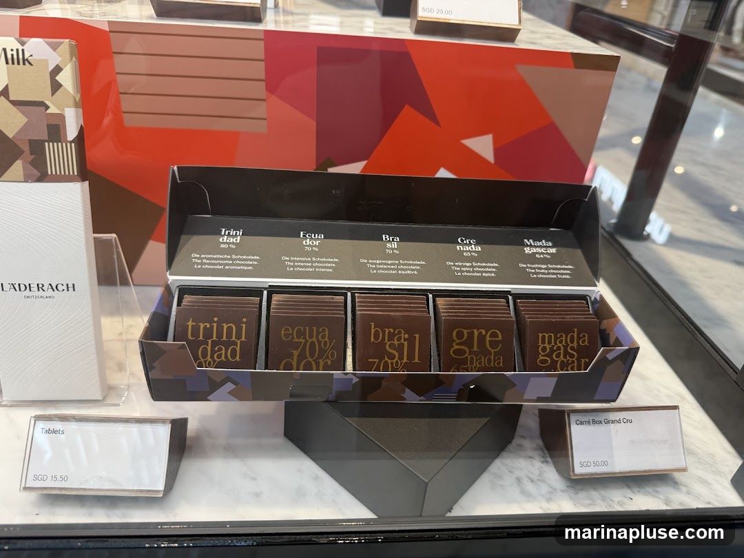 A display case shows an open box of assorted chocolates, with labels indicating origins and percentages. Individual chocolate squares are visible within the box. To the left, a white box with the brand name LÄDERACH and "SWITZERLAND" is displayed, along with a box labeled "Milk". Price tags for "Tablets" (SGD 15.50) and "Carré Box Grand Cru" (SGD 50.00) are also visible. A price tag of SGD 29.00 is also present.