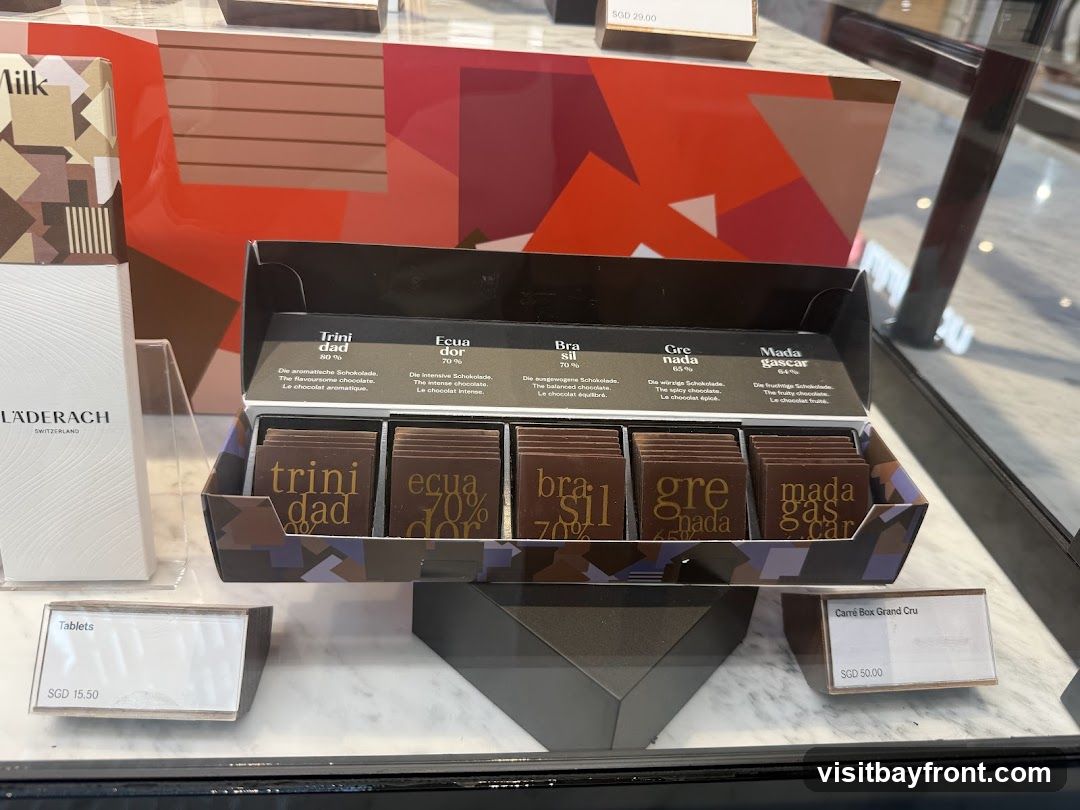 A display case shows an open box of assorted chocolates, with labels indicating origins and percentages. Individual chocolate squares are visible within the box. To the left, a white box with the brand name LÄDERACH and "SWITZERLAND" is displayed, along with a box labeled "Milk". Price tags for "Tablets" (SGD 15.50) and "Carré Box Grand Cru" (SGD 50.00) are also visible. A price tag of SGD 29.00 is also present.