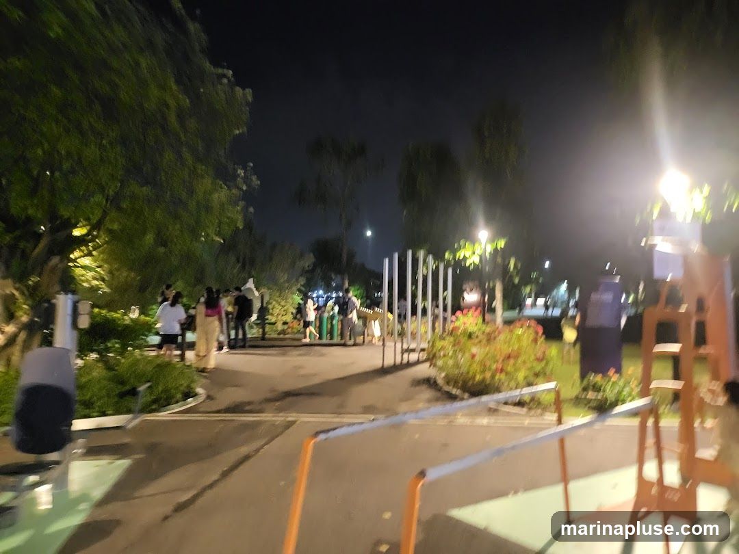 A nighttime view of a park with people walking along a path. There are trees, flower beds, and outdoor exercise equipment visible. Lights illuminate the area, creating a warm ambiance.