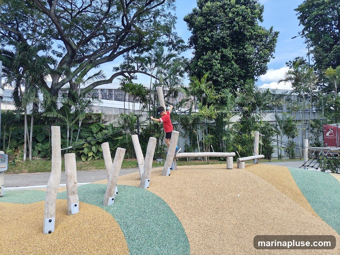 A young child in a red shirt and black shorts is navigating a rope obstacle course on a playground. The course consists of wooden posts and ropes, set against a backdrop of lush greenery and a blue sky. The playground surface is a mix of yellow and green rubberized material, designed to cushion falls. In the background, a red truck with a logo is partially visible.