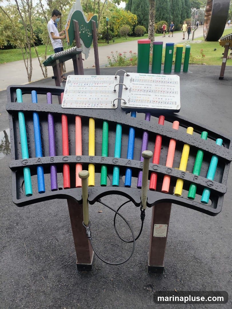 A colorful outdoor xylophone with sheet music displayed on a stand. In the background, a person plays a different musical instrument, and other people walk along a path in a park-like setting.