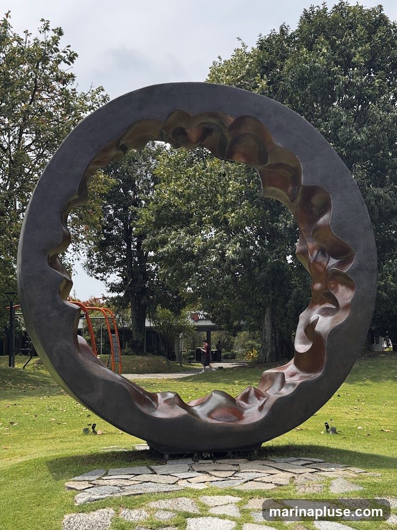 A large, abstract circular sculpture with an irregular, wavy interior is set in a grassy park. Trees surround the sculpture, and in the background, there is a building with a sign that appears to say "WC". A person is walking in the distance.