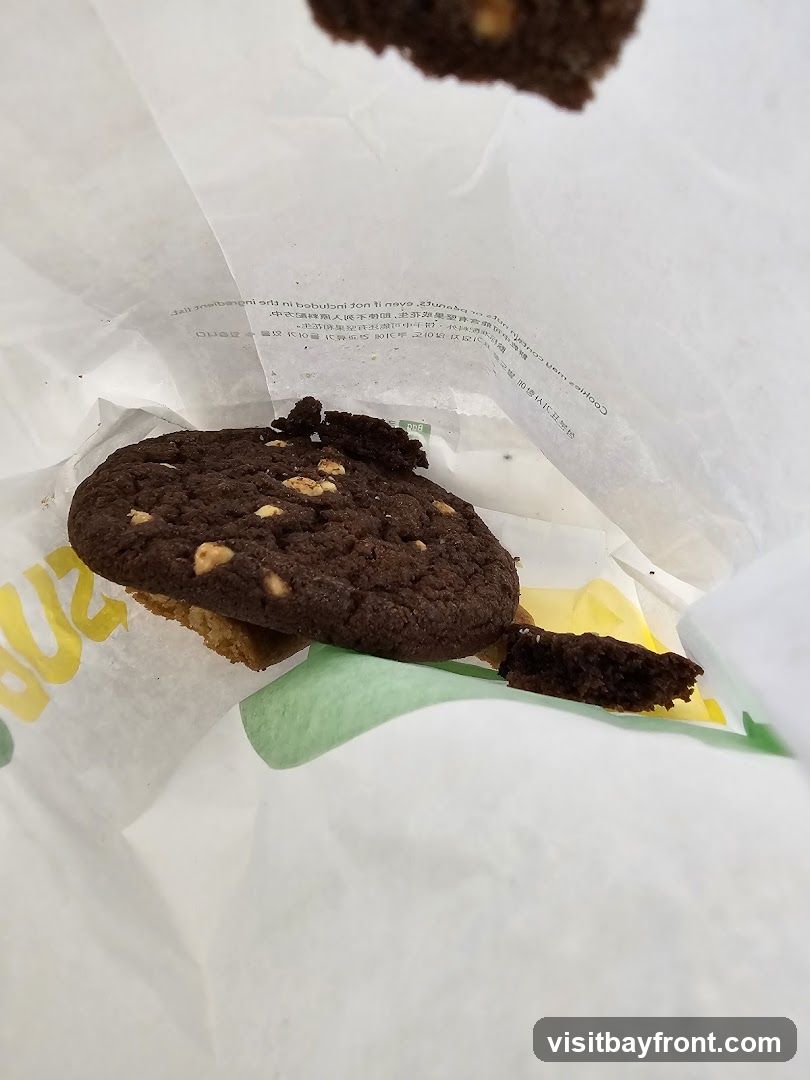 A close-up shot of a chocolate chip cookie with nuts, partially inside a white paper bag. The bag has some text printed on it, and a green and yellow design is visible. Another piece of cookie is seen in the foreground.