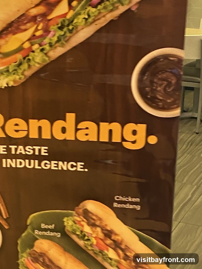A close-up of a promotional poster featuring sandwiches with rendang. The word "Rendang." is prominently displayed in large yellow letters. Below it, text reads "THE TASTE OF INDULGENCE.". Two sandwiches are pictured: one labeled "Beef Rendang" and another labeled "Chicken Rendang". A small bowl of dark sauce is visible in the upper right corner.
