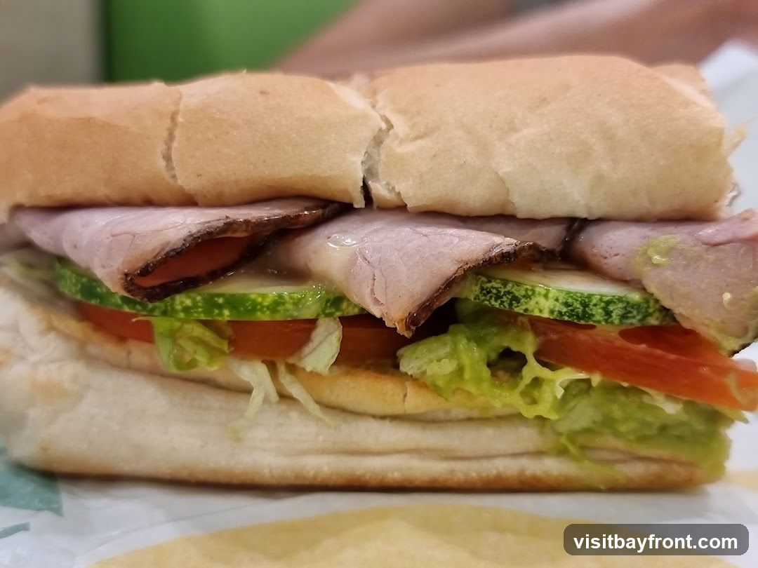 A close-up shot of a roast beef sandwich on a white paper wrapper. The sandwich is made with a long white bun, sliced roast beef, lettuce, tomato, and cucumber. Some green spread, possibly avocado or guacamole, is visible.