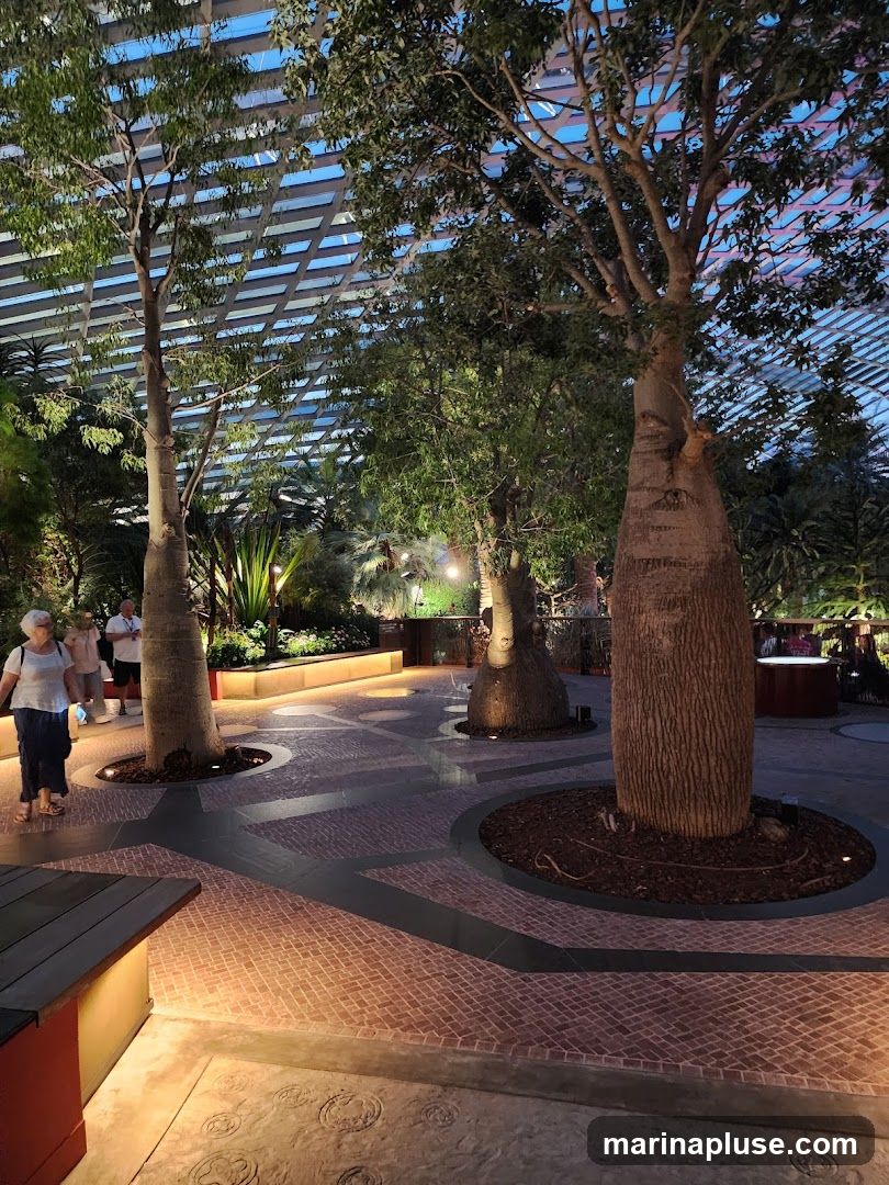 An indoor botanical garden with large trees, lush foliage, and a patterned floor. People are walking through the space, which is illuminated by soft lighting. The ceiling appears to be a glass or translucent structure with a grid pattern.