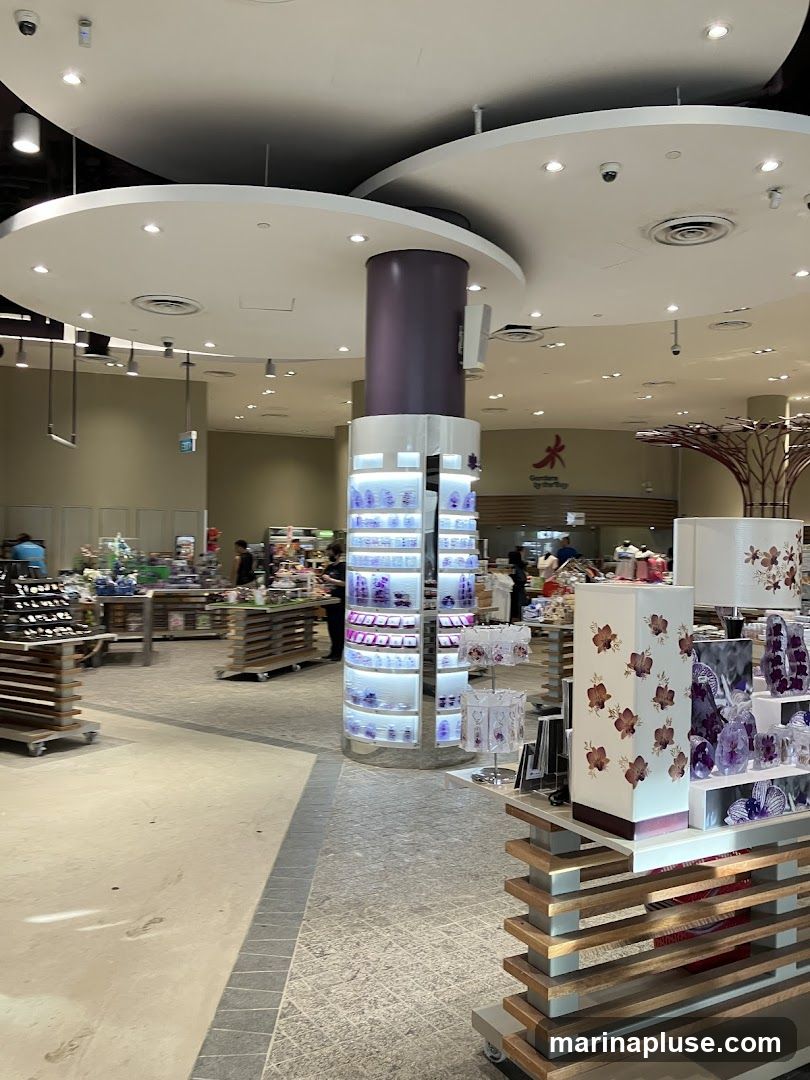 The image shows the interior of a well-lit retail store. In the foreground, there are display shelves with decorative items, including lamps and vases with floral patterns. A central cylindrical display unit showcases various products, possibly cosmetics or accessories, illuminated from within. In the background, more shelves and displays are visible, with a few customers browsing. A sign with a red flower logo and the text "Gardens by the Bay" is visible on the back wall.