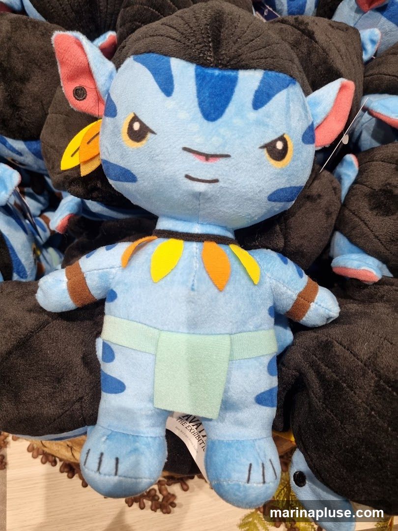 A close-up shot of a blue plush toy resembling a character from the movie Avatar. The toy has black hair, yellow eyes, and blue markings on its face and body. It is wearing a light green loincloth and a necklace of orange and yellow leaf-like shapes. The plush toy is surrounded by other similar toys, some of which are black and appear to be the backs of other Avatar characters. A tag is visible on the lower front of the toy, reading "AVATAR THE EXHIBITION" and "TM & © 2010 Century Studios".
