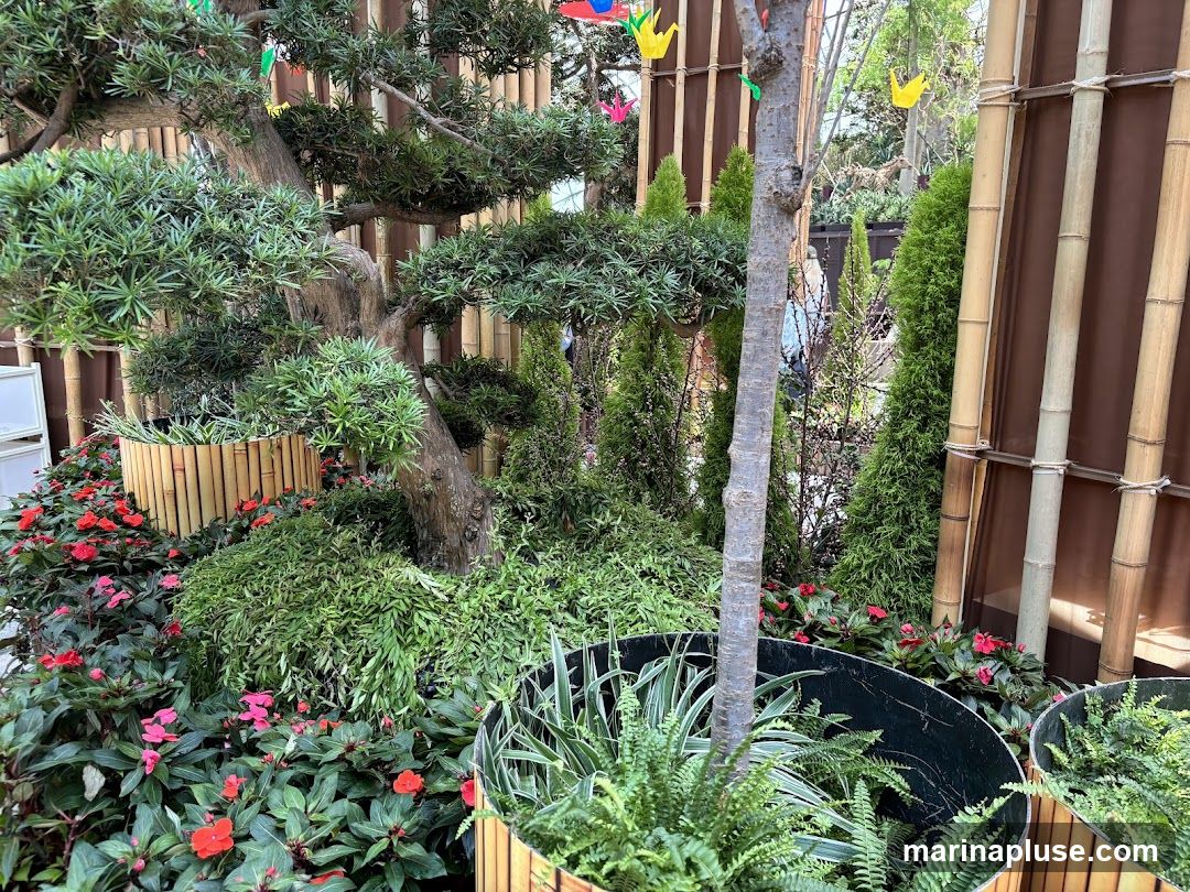 A lush garden display featuring a large bonsai tree, various green plants, and red and pink flowers. The background is decorated with bamboo poles and brown fabric. Small colorful origami cranes are hanging from above.
