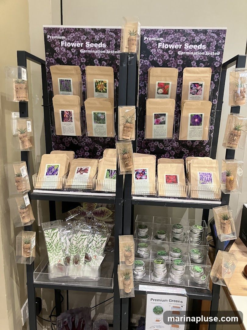 A retail display featuring various packets of flower seeds, small potted plants, and tins of greens. The display has multiple shelves with products organized in clear plastic containers and on shelves. Two large posters at the top advertise "Premium Flower Seeds, Germination tested."